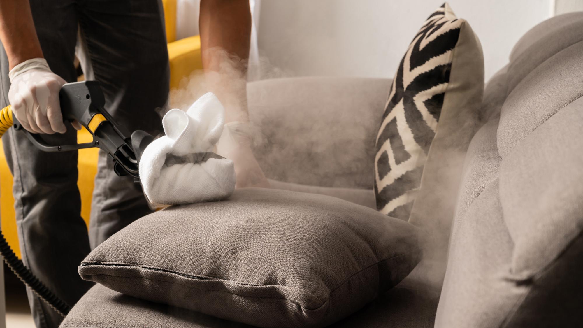 A person using a handheld steamer to clean a gray sofa, with steam visible around the cushion being cleaned; there is a black and white decorative pillow on the sofa.
